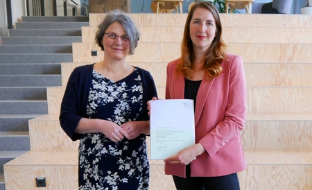 Prof. Dr. Annabella Rauscher-Scheibe & Dr. Kathrin Goldammer stehen vor einer Treppe. Dr. Kathrin Goldammer hält ihre Ernennungsurkunde in den Händen.