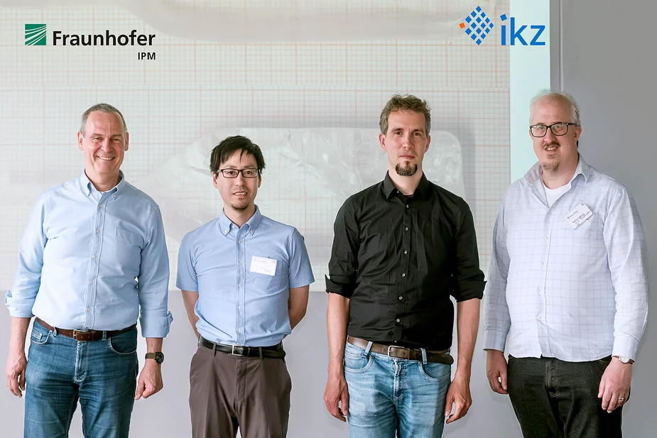 IKZ-employees in front of of an image of a BaMgF4 crystal grown at IKZ